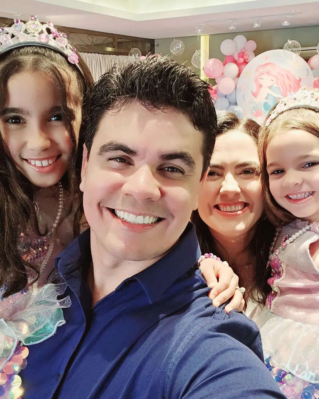 Rafael Santos sorrindo em selfie com a esposa e as duas filhas vestidas de princesas